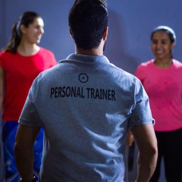 Back view of a personal trainer guiding two women during a gym session indoors.